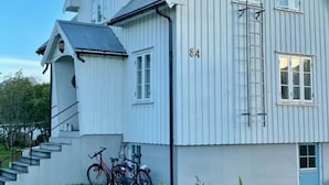 Une maison blanche à deux étages, avec un toit gris, un numéro « 84 » bien visible sur le côté et un vélo garé devant.