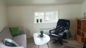 A living room with a grey sofa, a black leather chair, a white coffee table, and a window with potted plants.