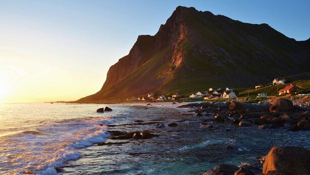 A coastal landscape with a prominent mountain, rocky shoreline, and small houses along the beach.