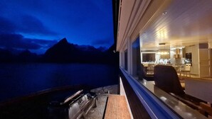 A modern interior with a kitchen and dining area visible through a window overlooking a body of water and mountains. - Lofoten panoramic luxury home with sauna (Moskenes)