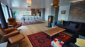 A modern living room with leather chairs, a sofa, a wooden coffee table, a red patterned rug, a stone fireplace, and a kitchen in the background.