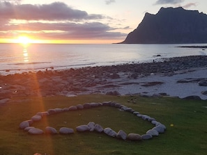 Un paysage côtier avec un coucher de soleil, une grande formation rocheuse et un cercle de pierres sur l’herbe.