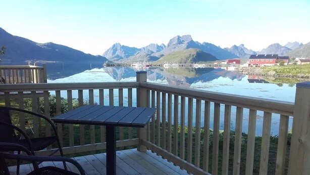 A wooden deck with a table and chairs, overlooking a calm lake with mountains in the background.