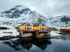 Una casa de madera con balcón sobre pilotes, sobre el agua, rodeada de montañas nevadas.
