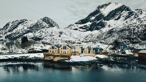 Una propiedad frente al agua con muelle, un edificio grande con balcón y montañas nevadas al fondo.