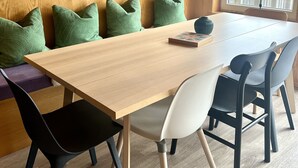 Un comedor con una mesa de madera, cojines verdes, sillas negras y blancas, una lámpara colgante transparente y un amplio ventanal con vista a un lago y montañas.