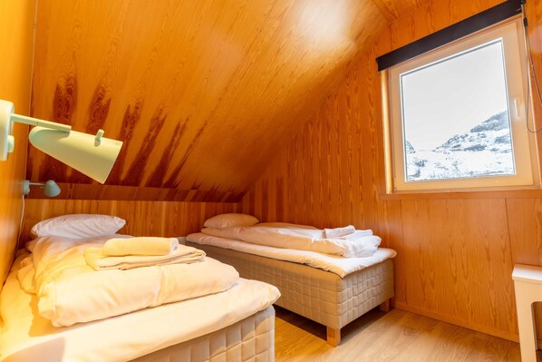 Una stanza in legno con due letti, una finestra che si affaccia su un paesaggio innevato e una lampada fissata al muro.
