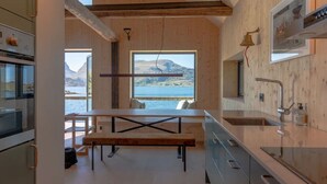 A modern kitchen with a wooden interior, a large window overlooking a body of water and mountains, and a dining area with a bench. - Minkfarmen: Spacious seaside cabin w/scenic view (Flakstad)