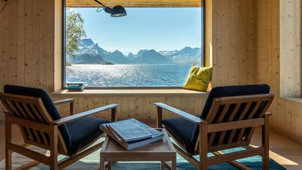 A room with wooden walls, a large window with a view of mountains and water, two chairs, a table with books, and a lamp. - Minkfarmen: Spacious seaside cabin w/scenic view (Flakstad)