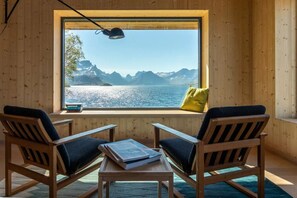 Une pièce aux murs en bois, avec une grande fenêtre donnant sur les montagnes et l’eau, deux chaises, une table avec des livres et une lampe.
