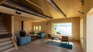 A wooden cabin interior with a staircase, a sofa, a table, and a large window overlooking a lake. - Minkfarmen: Spacious seaside cabin w/scenic view (Flakstad)