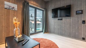 Habitació moderna amb parets de fusta, un televisor fixat a la paret, una porta corredissa de vidre que dona a un balcó, una taula petita amb una espelma i un comandament a distància, i una catifa vermella.