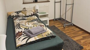 Un lit simple avec une couvre-lit à motifs floraux, une table de chevet avec une lampe, un radiateur, une fenêtre avec des stores et un étendoir métallique.