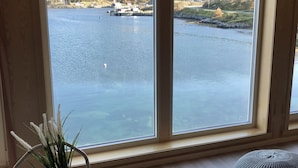 A room with a large window overlooking a lake and mountains, a small table with a plant, and two grey poufs.