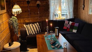 A cozy log cabin interior with a wooden floor, a sofa, a coffee table, a lamp, and a window with curtains.