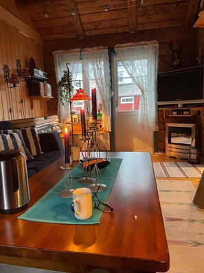 A cozy living room with a wooden table, a sofa, a television, and a window with curtains. - Koselig rorbu på Tind (Moskenes)