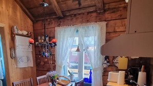 A wooden cabin interior with a dining area, kitchen, and a window with lace curtains.
