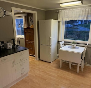 Uma cozinha compacta com geladeira, mesa de jantar com cadeiras, relógio e janela com vista.
