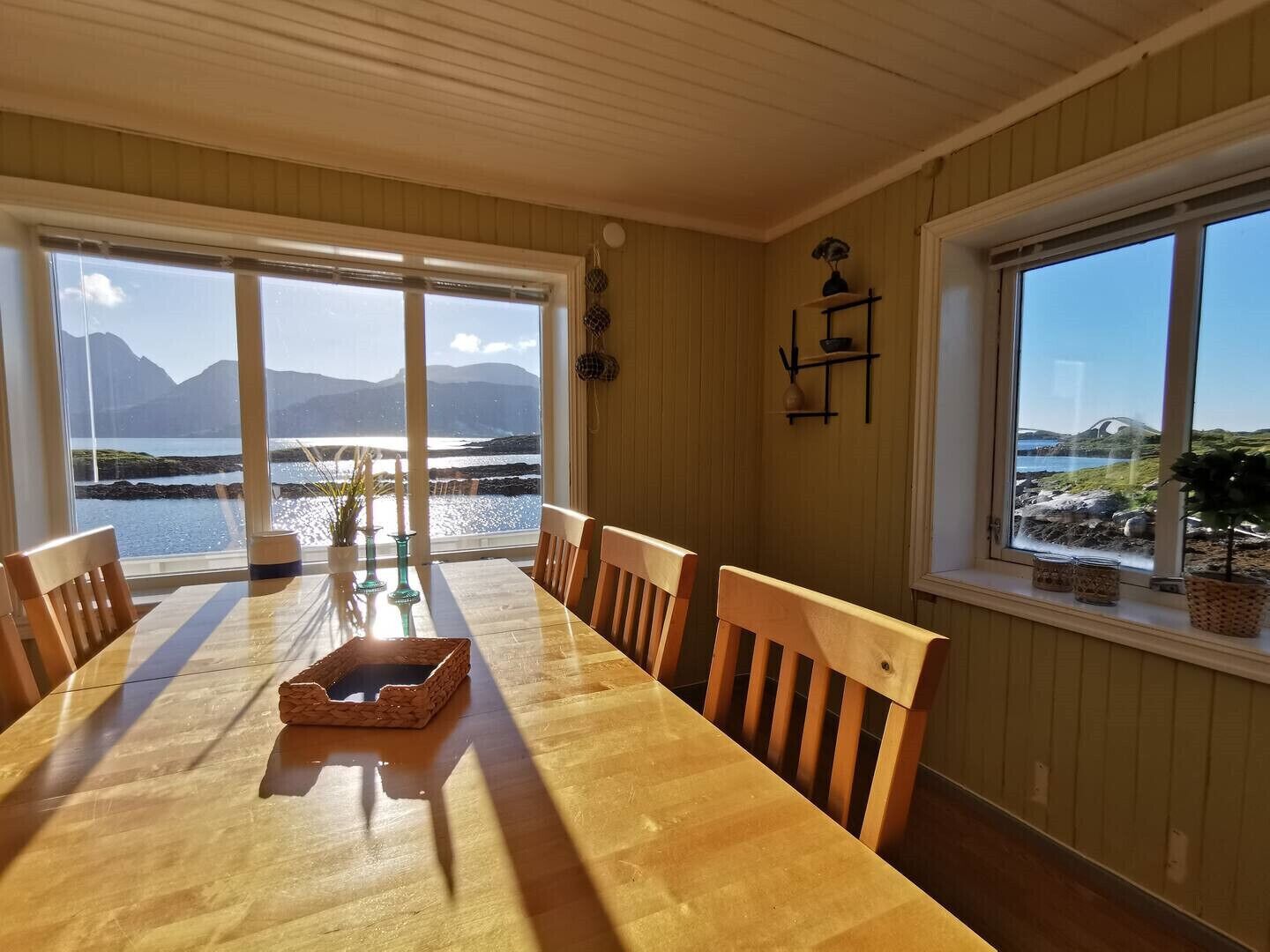 A dining area with a wooden table and chairs, large windows with a view of a body of water and mountains, and a wall-mounted shelf with decorative items.