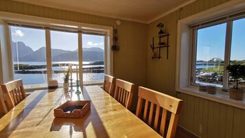 A dining area with a wooden table and chairs, large windows with a view of a body of water and mountains, and a wall-mounted shelf with decorative items.