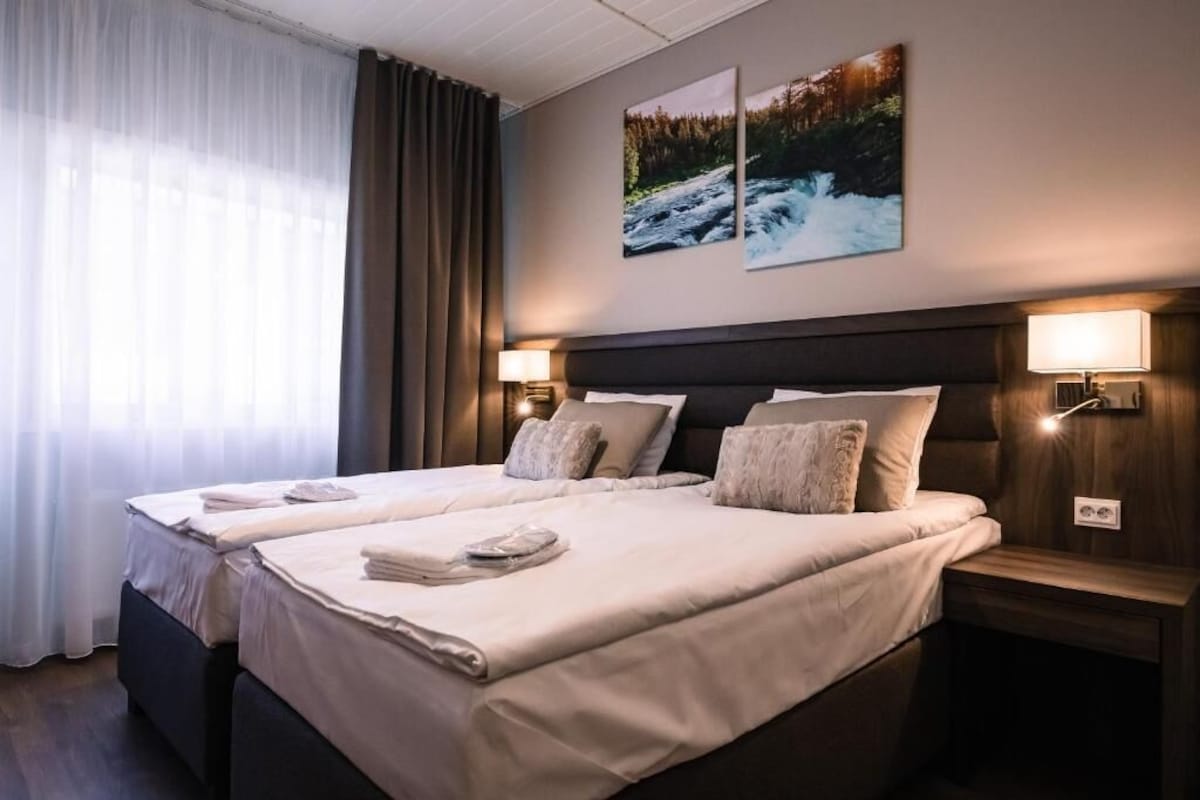 A hotel room with two beds, a large window with curtains, a wall-mounted picture, and bedside lamps.