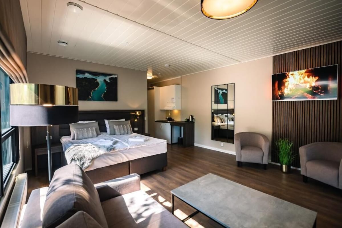 A modern hotel room with a bed, sofa, armchair, desk, and a view of the sea.