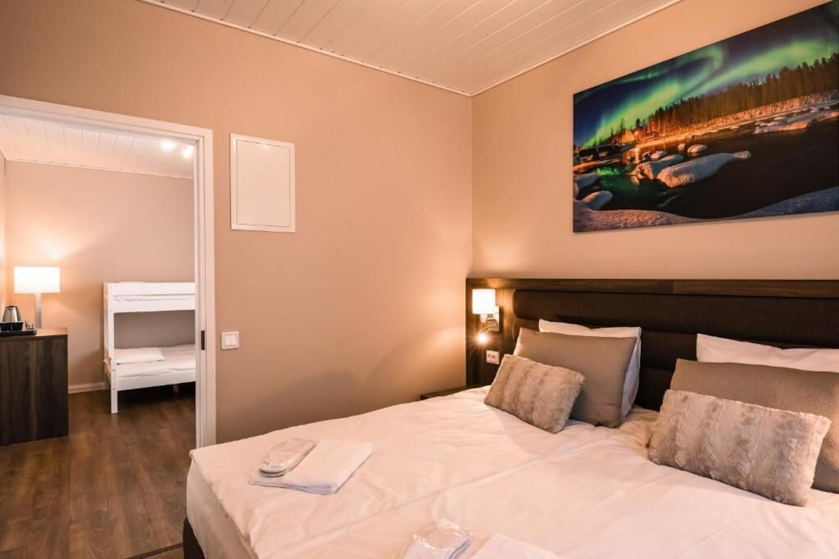 A hotel room with a bed, a nightstand, a lamp, a painting of the Northern Lights, and a bunk bed in the adjacent room.