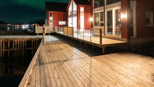 Une jetée en bois mène à un bâtiment rouge doté de grandes fenêtres et d’une porte en verre.