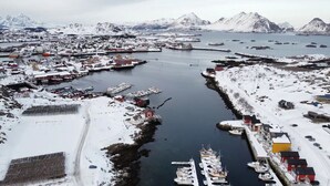 Une ville côtière aux bâtiments recouverts de neige, un port de plaisance avec des bateaux, et des montagnes en arrière-plan.