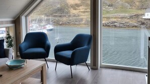 Une pièce moderne avec deux fauteuils bleus, une table en bois et une grande fenêtre donnant sur un plan d’eau et des collines.