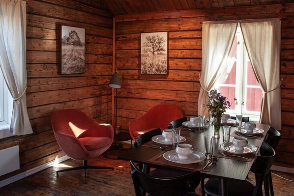 A wooden cabin interior with a dining table set for four, two red chairs, a vase with flowers, and two framed pictures on the walls.