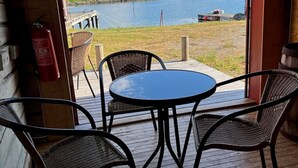 A wooden deck with a round table and four wicker chairs, overlooking a body of water with a dock and buildings in the distance. - Tradisjonell Rorbu i fiskeværet Ballstad (Vestvågøy)