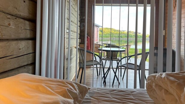 A cabin room with a bed, a table with chairs, a window with blinds, and a view of a lake and greenery outside. - Tradisjonell Rorbu i fiskeværet Ballstad (Vestvågøy)