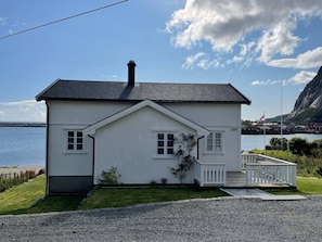 A white two-story house with a black roof, a small porch, and a view of the sea. - Nordlandshus ved havet på Reine, Lofoten (Moskenes)