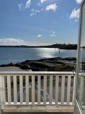 Vista da un balcone con ringhiera, su un tratto d'acqua tranquillo, una costa rocciosa e edifici in lontananza.