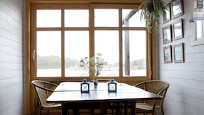 Une table à manger en bois, accompagnée de quatre chaises, d’une plante en pot et offrant une vue sur un plan d’eau grâce à de grandes fenêtres.