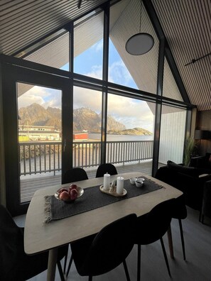 Un coin repas avec une table dressée pour deux personnes, de grandes fenêtres offrant une vue sur les montagnes et un plan d’eau, et un balcon doté d’une balustrade.