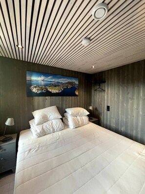 A modern bedroom with a wooden ceiling, a large bed with white bedding, a nightstand with a lamp, and a wall-mounted painting. - Rorbuleilighet i toppetasjen i Henningsvær (Vågan)