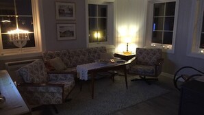Ein Wohnzimmer mit einer Couch, einem Sessel und einem Tisch mit Lampe. An der Wand hängen Bilderrahmen, und ein Fenster bietet einen Ausblick.