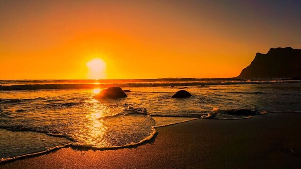 Ein Sonnenuntergang über einem Strand mit Wellen und einer in der Ferne liegenden Klippe.