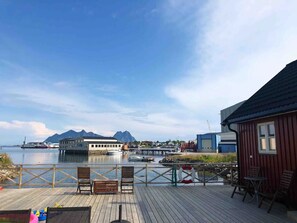A wooden deck with chairs and tables, a lifebuoy, and a view of the water and mountains.