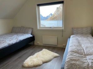 A bedroom with two beds, a window with a view of mountains, a radiator, and a fur rug on the floor. - House in Reine by the Fjord (Moskenes)