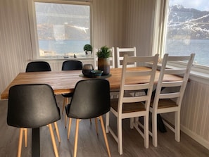 A dining area with a wooden table, black chairs, and a view of a mountainous landscape through a window. - House in Reine by the Fjord (Moskenes)