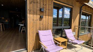 Une façade en bois avec un balcon, deux fauteuils violets rembourrés, une petite table en bois et une porte vitrée donnant sur un espace intérieur.