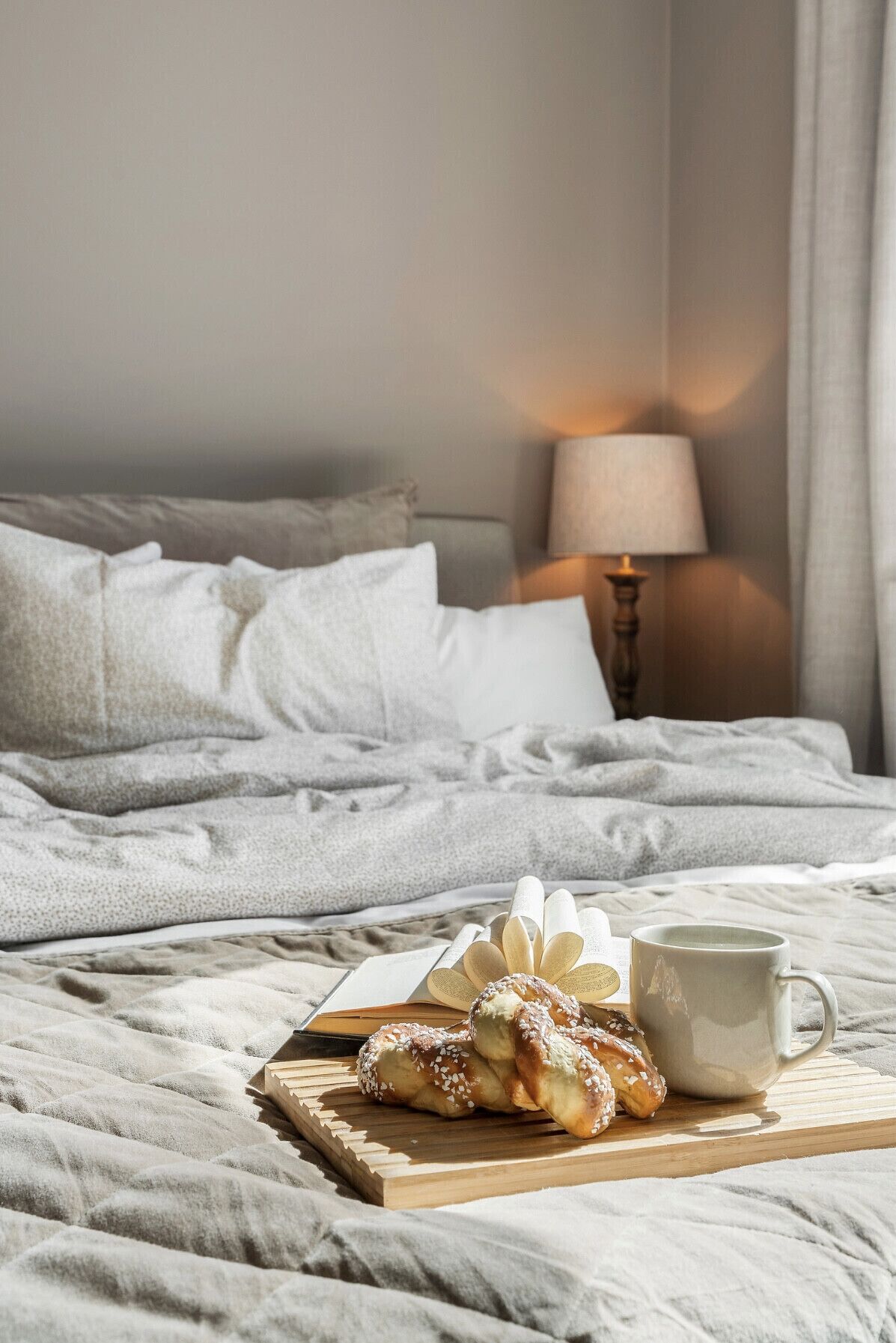 Ein ordentlich bezogenes Bett mit Kissen, einer Nachttischlampe und einem Tablett mit Gebäck und einer Tasse auf einer Holzbrett.