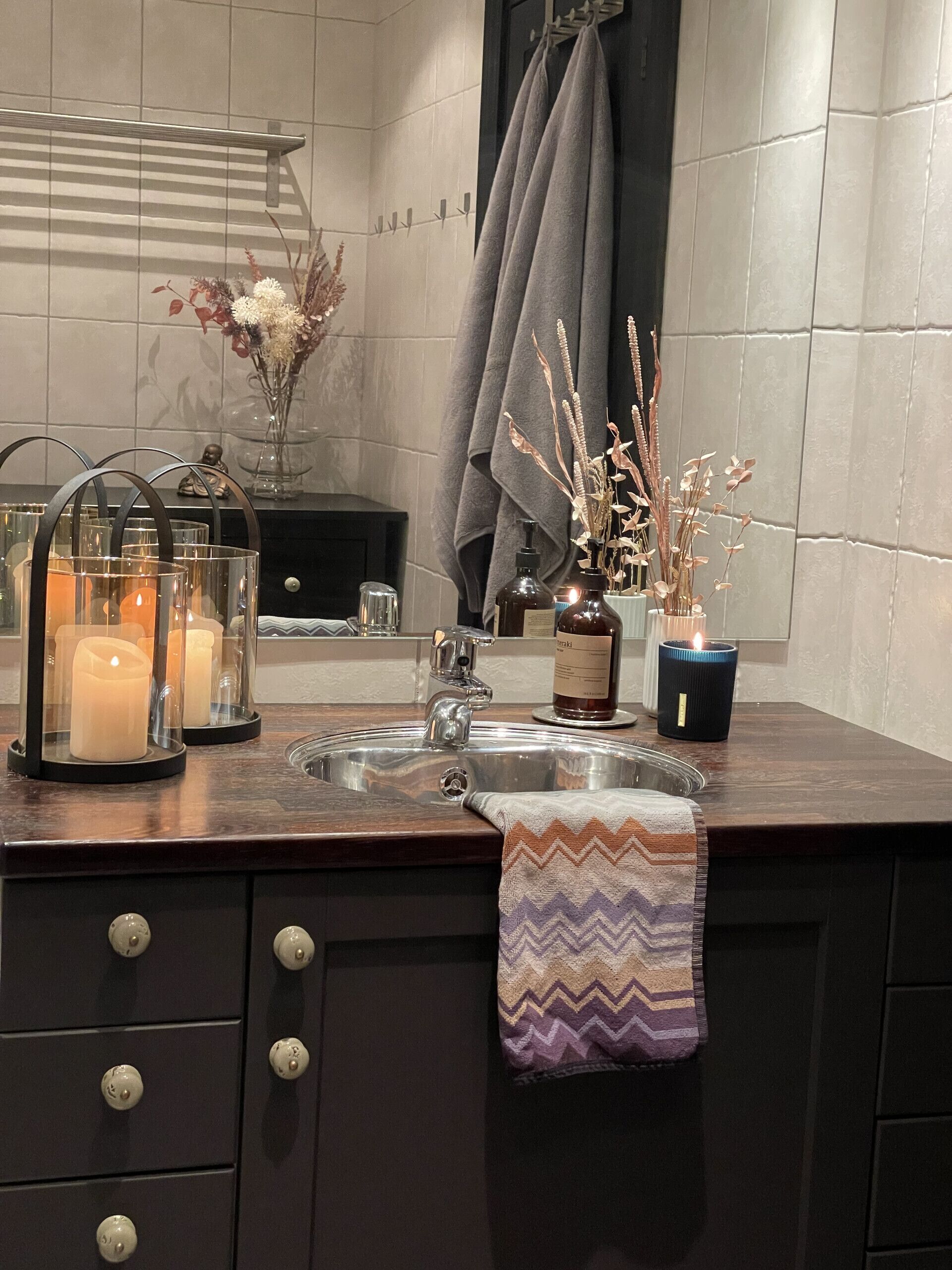 A bathroom with a sink, mirror, towel rack, and decorative items including candles and a vase with dried plants.
