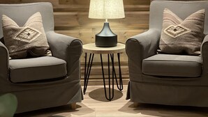 Una habitación con pared de madera, dos sillones, una mesa con lámpara y un cuadro de un sendero nevado en la montaña.