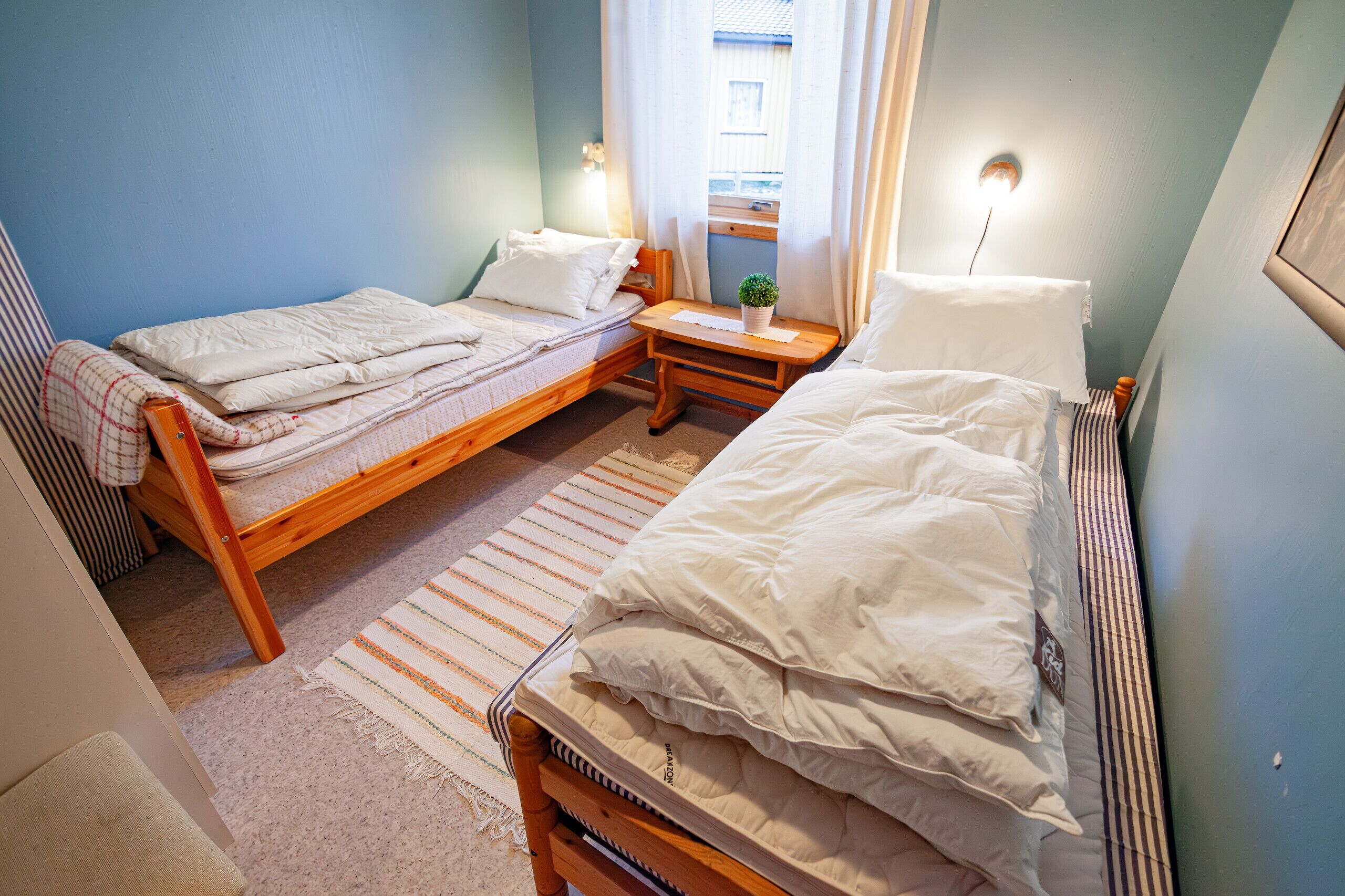 Quarto pequeno e acolhedor com duas camas de solteiro, uma mesinha de cabeceira de madeira, uma pequena mesa com uma planta e uma janela com cortinas.