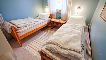 Quarto pequeno e acolhedor com duas camas de solteiro, uma mesinha de cabeceira de madeira, uma pequena mesa com uma planta e uma janela com cortinas.