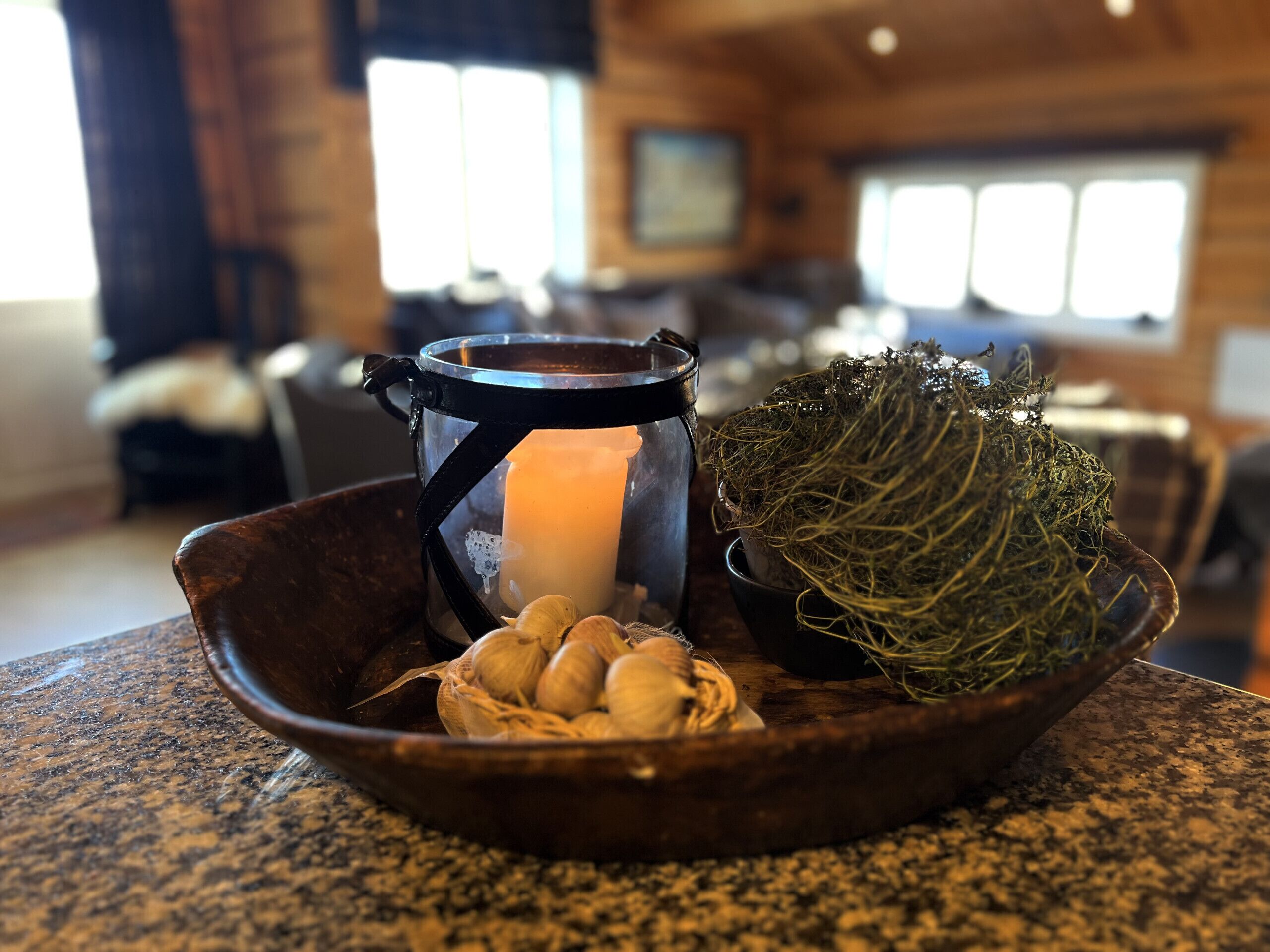 A candle in a glass jar, garlic bulbs, and a moss ball on a wooden table in a room with a kitchen in the background.